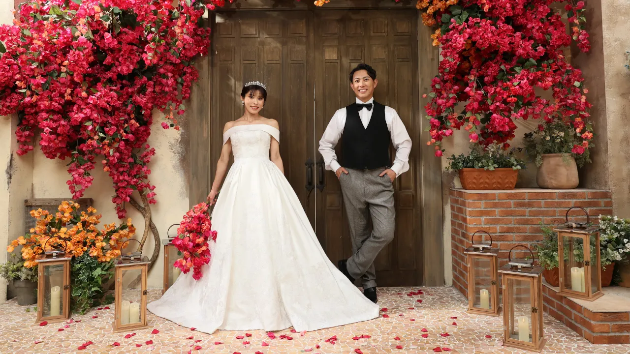 白いウェディングドレスとタキシード姿のカップルが花に囲まれた扉の前でポーズ